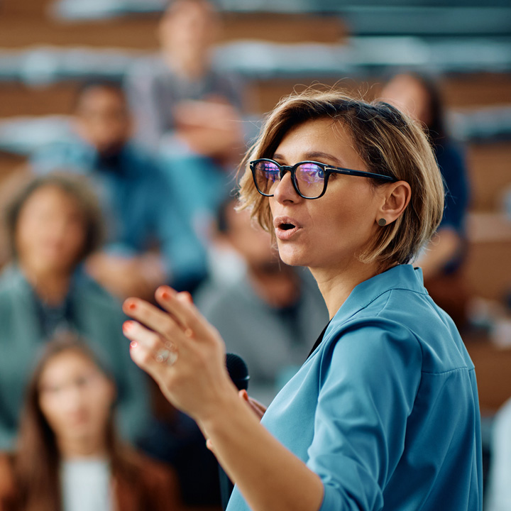 Woman wearing glasses, presenting to a room of people